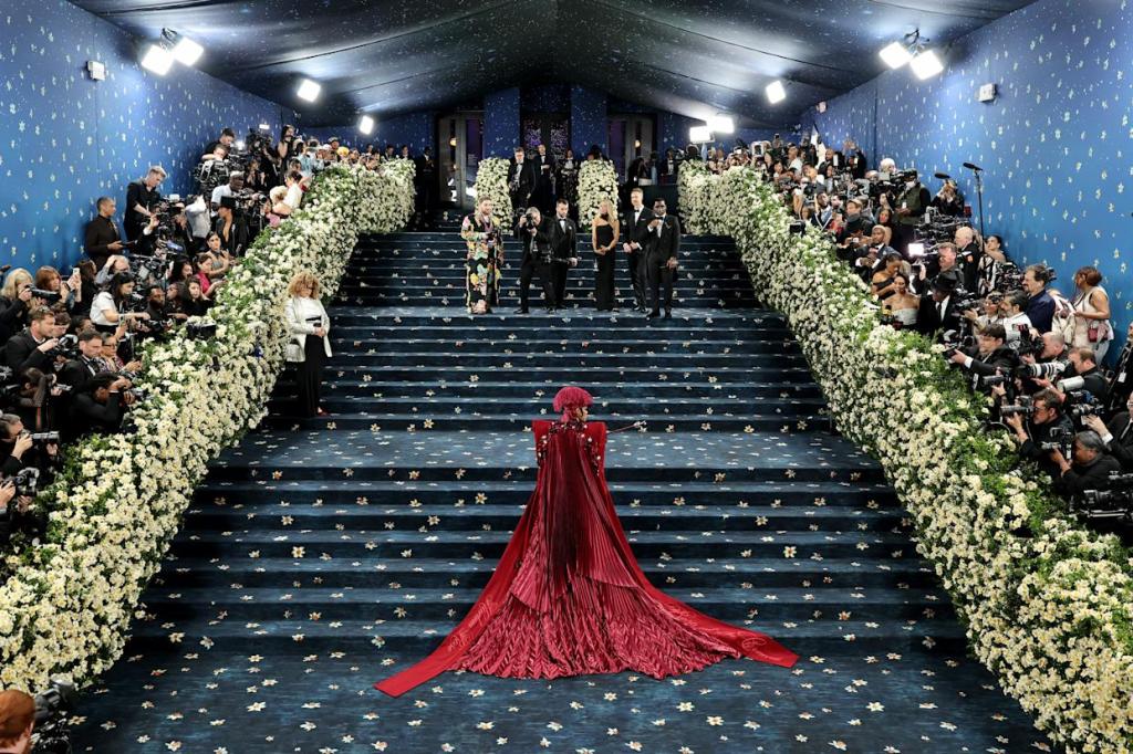 Teyana Taylor at the Met Gala Teyana Taylor on the steps of the Met Gala dressed in custom Marc Jacobs, and styled by Ruth E. Carter. She is wearing a dark red cape and a hat with a feather, and the steps have a blue carpet with flowers on, and flower bushes around filled with paparazzis.
